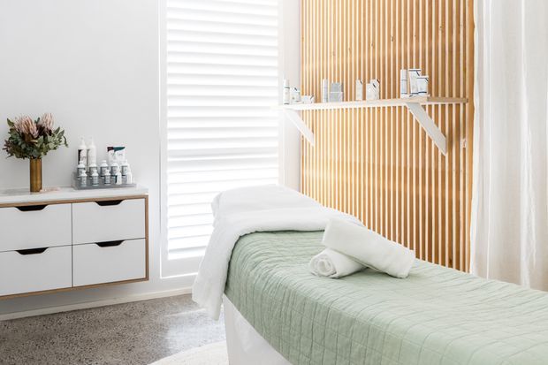 Bright, serene treatment room with a massage table dressed in pale green and white linens, a white floating storage cabinet, spa products, dried floral arrangement, white plantation shutters, and a timber slatted feature wall with a simple display shelf.