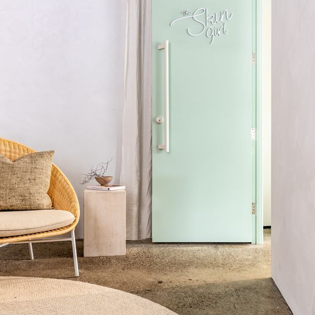 A softly styled interior featuring a mint-green door with a long vertical handle and decorative lettering, opening into another room. A woven rattan lounge chair with neutral cushions sits beside a small pale timber side table, with curtains, textured walls, and a muted carpet creating a warm, relaxed setting.