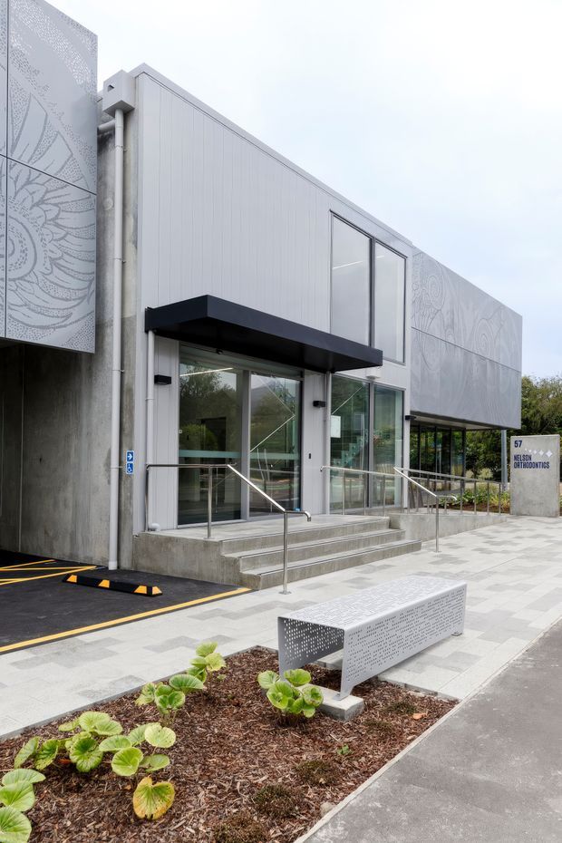 Exterior view of a contemporary commercial building entrance with pale vertically profiled cladding, extensive glazing, a black projecting canopy, concrete steps, stainless steel handrails, and accessible ramp access. Decorative perforated metal panels with botanical patterns frame the facade. A paved forecourt includes a perforated metal bench, planted garden beds, signage, and marked parking.