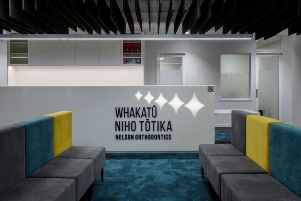 A contemporary orthodontic clinic reception or waiting area with symmetrical grey upholstered bench seating, teal and yellow accent panels, a blue patterned carpet, white built-in cabinetry, frosted-glass doors, and prominent bilingual clinic signage. A dramatic black slatted ceiling adds contrast, while geometric light projections illuminate the rear wall.