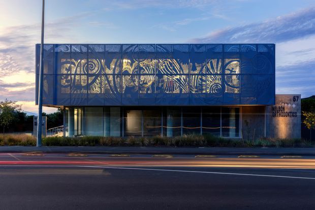 Street-facing view of a contemporary commercial building at dusk, featuring a dark perforated or patterned upper facade with illuminated graphic detailing, extensive ground-floor glazing, and a landscaped frontage beside a roadway. A vertical sign identifies the premises as Nelson Orthodontics.