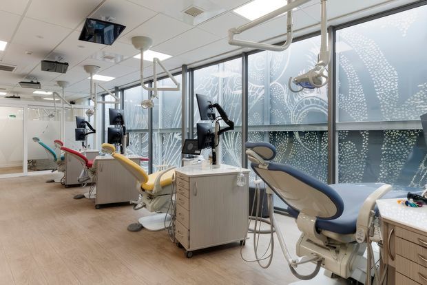 A bright contemporary dental clinic treatment room with several dental chairs, mobile cabinetry, overhead examination lights, monitors, and exposed service piping. Floor-to-ceiling windows feature patterned privacy film, while pale timber-look flooring and light neutral finishes create a clean, airy clinical environment.