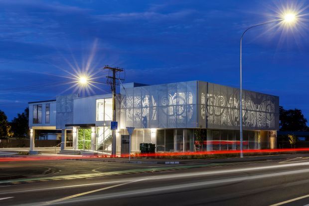 Night-time exterior view of a contemporary two-storey commercial building on a busy urban street. The building has a translucent, patterned metal facade with illuminated interiors, extensive glazing, a projecting upper volume, and strong street lighting with long-exposure traffic light trails.