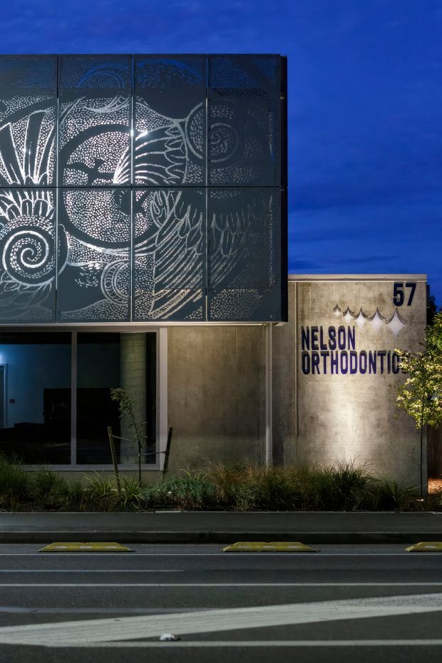 Night-time exterior view of a contemporary orthodontic clinic. The facade combines a dark patterned perforated metal screen with abstract botanical artwork, smooth concrete walls, extensive dark-framed glazing, and illuminated signage reading “Nelson Orthodontics” with the number 57. A landscaped strip and road occupy the foreground.