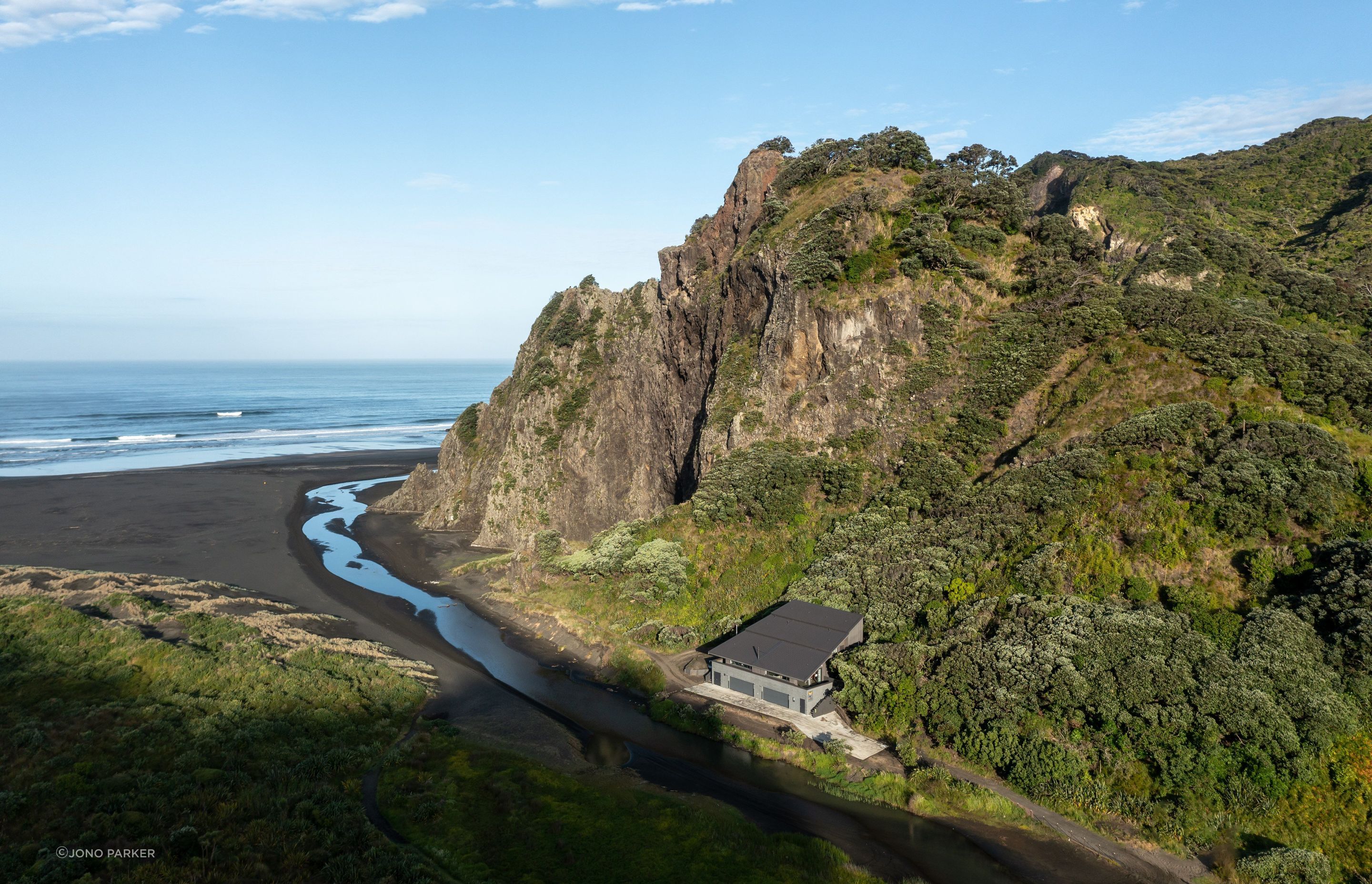 ©️ Jono Parker     Nestled into the base of The Watchman basalt cliffs, the Trusts Karekare Surf Life Saving Club takes in the same earthy hues.