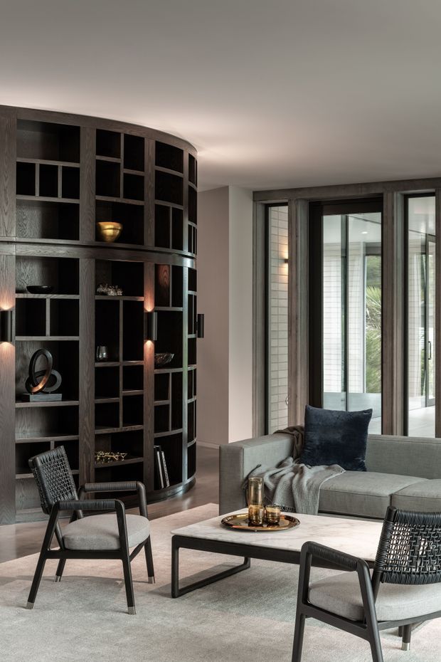 A refined contemporary living room with a large curved dark timber built-in shelving unit, soft grey sectional sofa, black-framed lounge chairs, pale stone coffee table, and layered neutral furnishings. Full-height glazed doors and windows bring in natural light, while integrated warm lighting highlights the shelving and decorative objects.