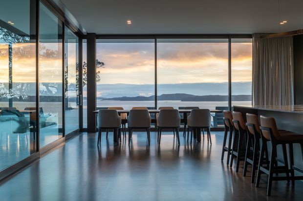 A contemporary open-plan dining and entertaining space with floor-to-ceiling glazing overlooking a calm coastal or waterfront landscape at sunset. A long dining table with upholstered chairs sits centrally, while a substantial island or bar with high stools occupies the right side. The room features dark-framed glazing, polished concrete flooring, a dark ceiling, and full-height curtains, creating a refined, spacious interior with strong indoor-outdoor connection.