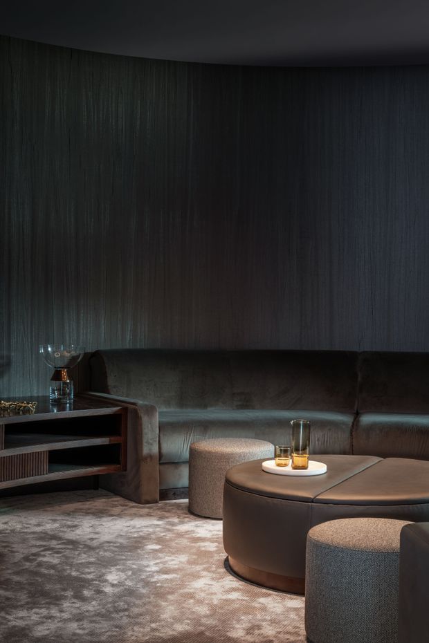 A dark, sophisticated lounge with a curved black-brown upholstered sectional sofa, rounded ottomans, and a large low circular coffee table. The walls are clad in vertically textured dark timber or wood-look panels, with a dark ceiling, low ambient lighting, and a soft patterned grey-brown rug. Built-in open shelving and a small drinks arrangement reinforce the intimate, luxurious atmosphere.