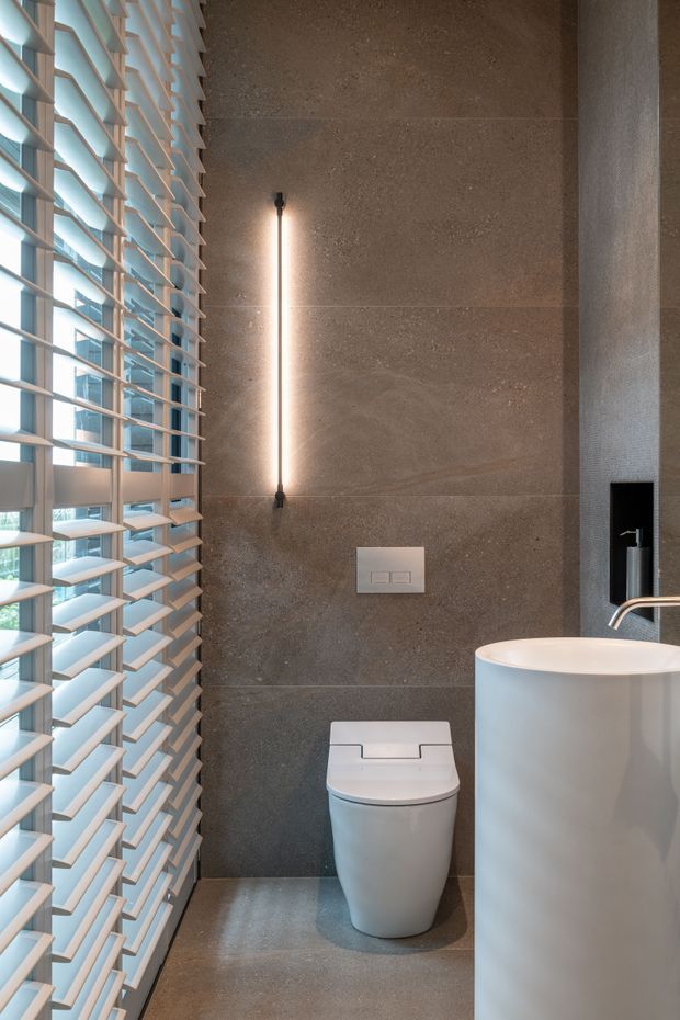 A compact contemporary bathroom featuring large-format dark grey stone-look wall and floor surfaces, a white wall-mounted toilet, and a rounded white basin with brushed metal tapware. Full-height louvred window shutters bring in daylight along the left side, while a slim vertical wall light adds a warm accent. A black recessed fixture and flush plate provide contrast.