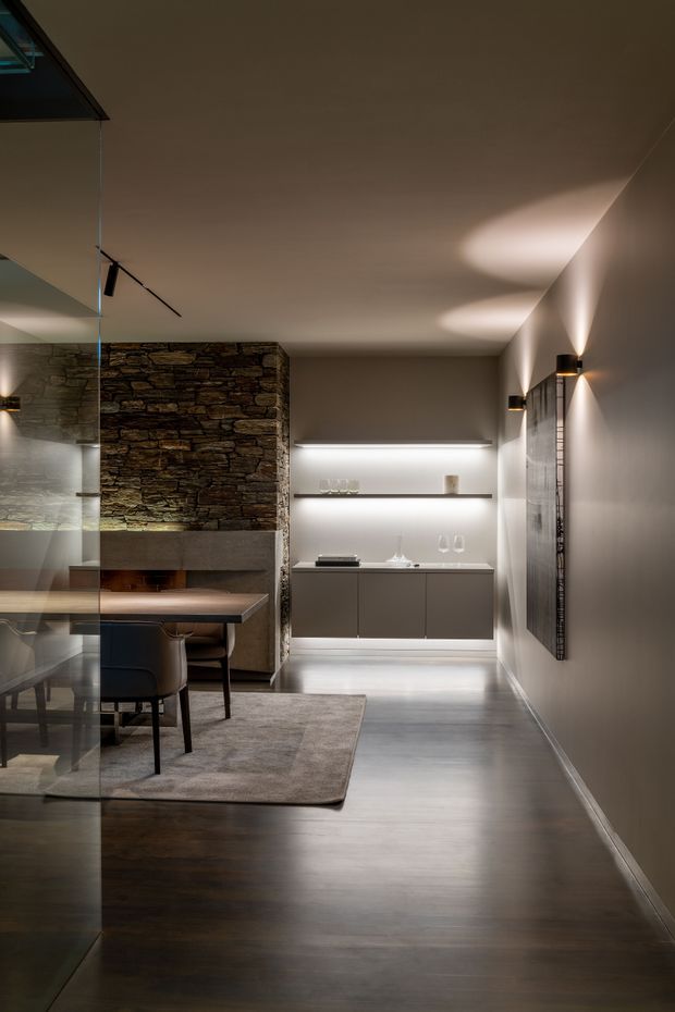A refined contemporary dining and living area with a dark timber floor, neutral walls, recessed linear shelving and floating cabinetry. A substantial stacked-stone fireplace wall forms the main feature, while a glass partition reflects the dining table and upholstered chairs. Discreet wall sconces and concealed shelf lighting create a warm, atmospheric evening ambience.