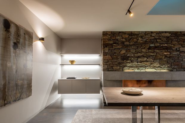 A refined contemporary living and dining area with a long timber dining table, sculptural pendant lighting, built-in shelving, a dramatic stacked-stone feature wall, and an integrated fireplace with a concrete surround. The space uses warm indirect lighting, neutral walls, dark cabinetry, and polished grey flooring, with artwork and restrained styling creating a calm, sophisticated atmosphere.
