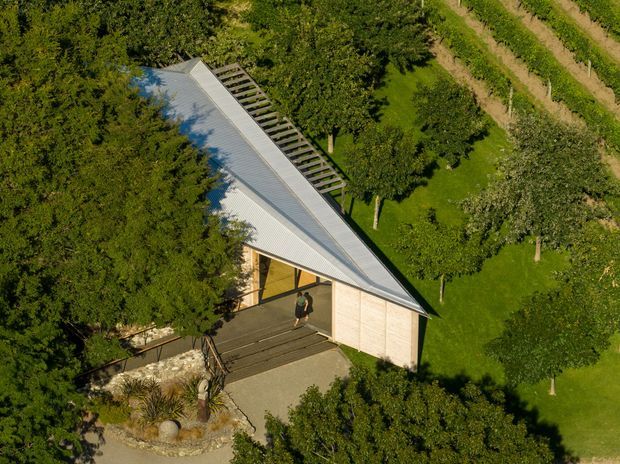 Aerial view of a compact contemporary building set within a lush landscaped rural or orchard-like property. The structure has a sharply angular single-slope metal roof, pale timber or light-coloured cladding, a covered entry, and a long open pergola or screened structure along the roof edge. Paved paths and planted garden beds surround the building.