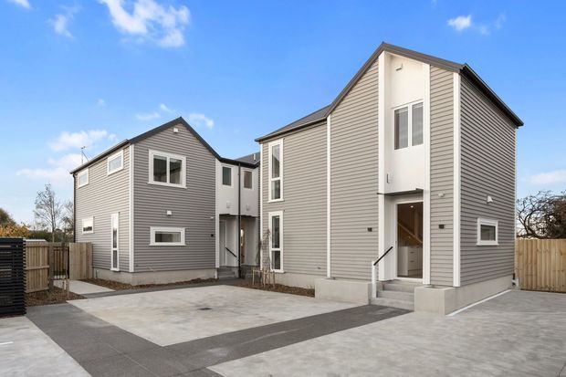 Exterior view of a contemporary multi-unit residential development with two-storey grey horizontal cladding, steep gabled rooflines, white-trimmed windows, recessed entry doors, and a broad concrete-paved courtyard or driveway area enclosed by timber fencing.