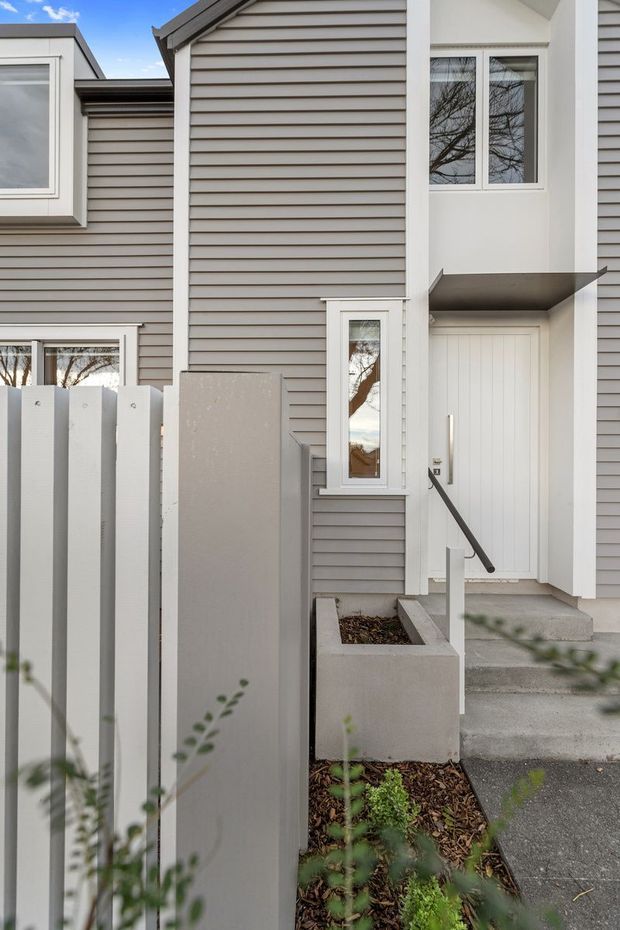 Exterior view of a contemporary multi-level townhouse entry. The facade is clad in horizontal grey weatherboards with crisp white trim, a recessed white panelled front door, narrow windows, concrete steps, and a small raised planter beside a slatted privacy fence.