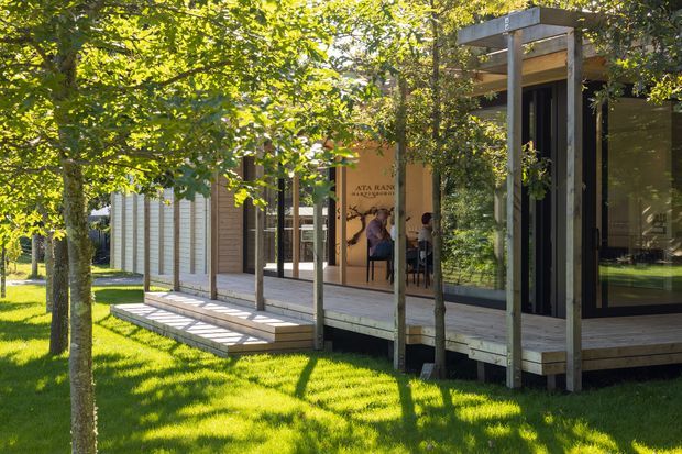 Exterior view of a contemporary single-storey pavilion-like building set within a leafy landscaped site. The structure features extensive floor-to-ceiling glazing, a raised timber deck and stepped access, vertical timber posts, pale horizontal wall cladding, and a flat pergola-like roof edge. Interior seating and signage are visible through the glazing, but the exterior terrace and building envelope dominate the image.
