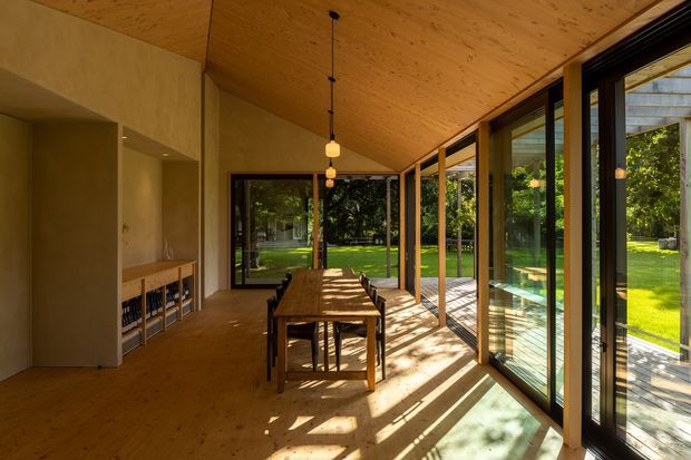 Warm, light-filled dining area with a long timber dining table and chairs, set within a contemporary glass-walled pavilion. The room features extensive floor-to-ceiling glazing, a timber-lined sloping ceiling, timber flooring, dark-framed sliding glass doors, and built-in shelving or cabinetry along the left wall. Strong sunlight creates long shadows across the floor, with a landscaped garden visible outside.