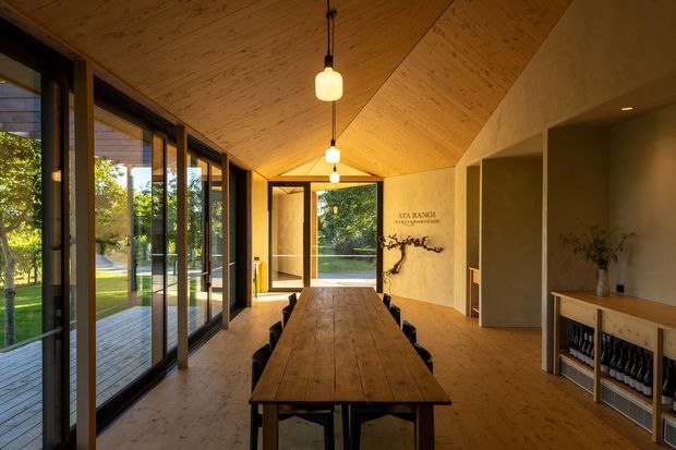 Warm, elongated dining room with a dramatic pitched timber ceiling and matching timber flooring. A long rustic timber dining table is centered beneath a row of pendant lights, with extensive dark-framed glazing and sliding doors opening toward a landscaped garden. The room also includes a built-in sideboard and shelving, textured plaster walls, and a sculptural wall feature, creating a refined natural contemporary atmosphere.