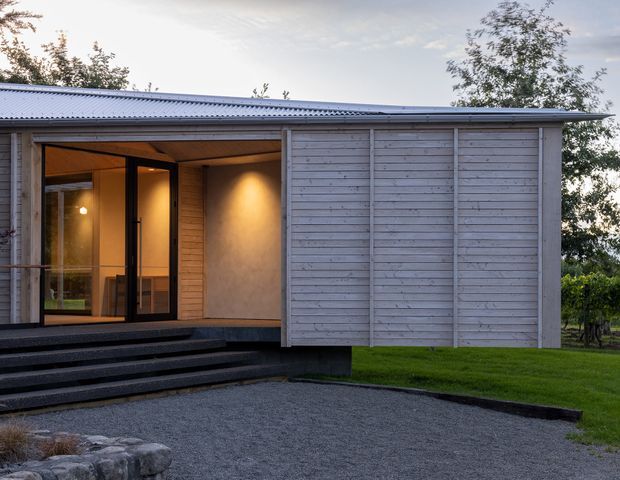 Single-storey contemporary building viewed from the exterior, with a low-pitched corrugated metal roof, horizontal pale timber cladding, a deep covered entry terrace, dark-framed glazed doors, and broad dark steps leading to a gravel forecourt. Warm interior lighting is visible through the glazing, with lawn and trees surrounding the structure.