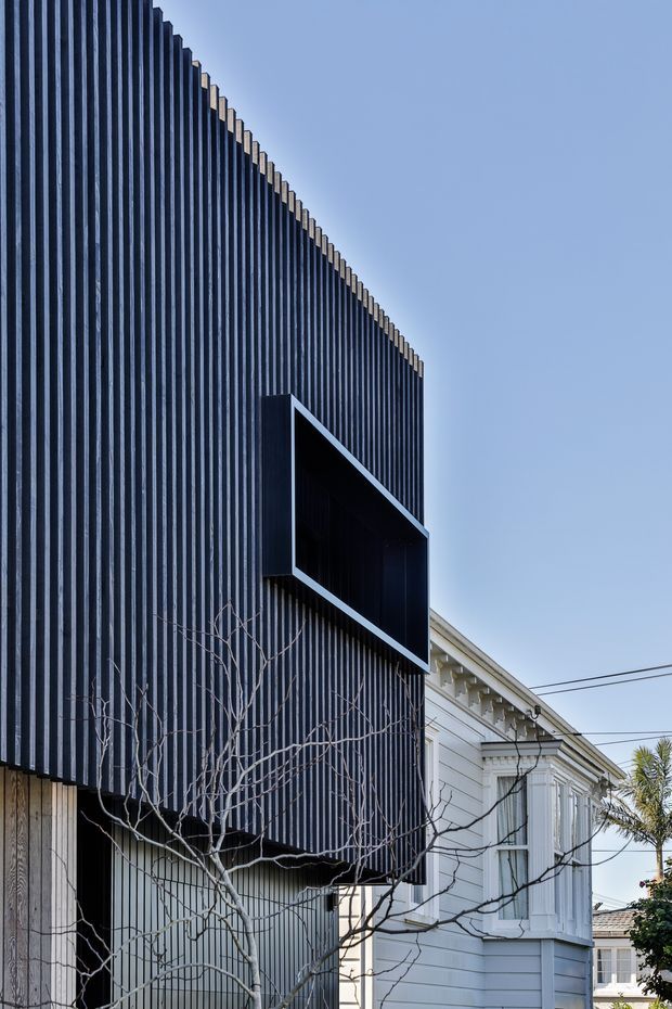Exterior architectural photograph showing a contemporary dark vertically ribbed facade and projecting box-like feature over a recessed opening, adjoining a preserved white weatherboard heritage-style building. Clear blue sky and overhead utility wires are visible; the image is dominated by the building envelope and its contrast between modern and traditional forms.