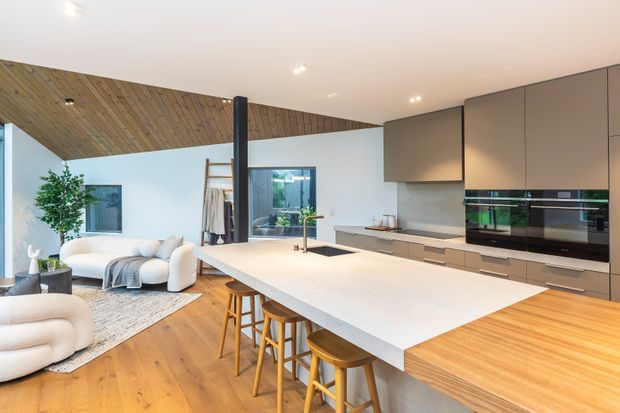 Open-plan contemporary kitchen and living area with a large white island, timber stools, integrated dark cabinetry and appliances, light timber flooring, and a warm timber-lined angled ceiling. A relaxed seating area and indoor greenery are visible beyond the kitchen.
