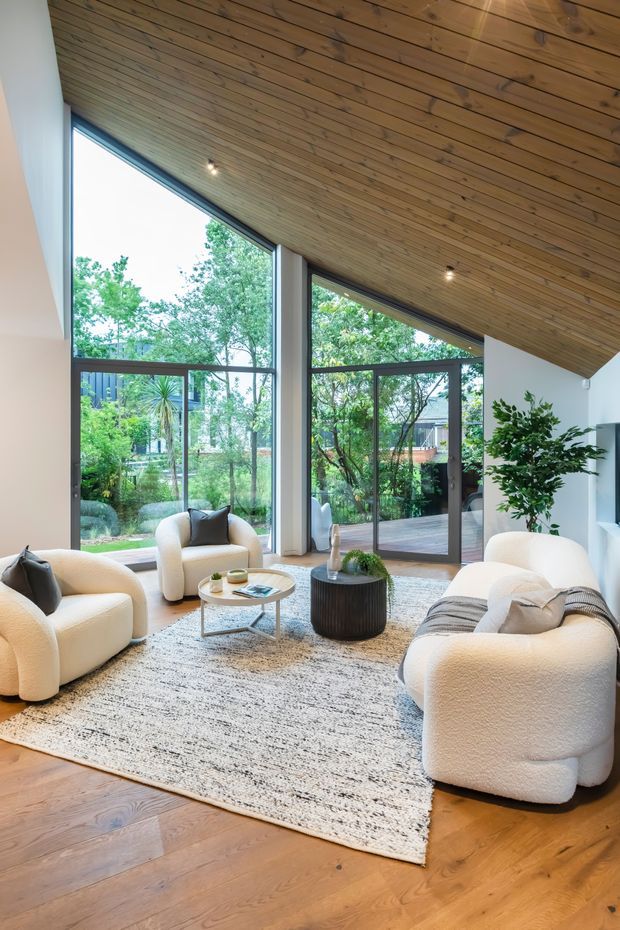 A bright, spacious living room with large floor-to-ceiling glazed openings overlooking a leafy garden and deck. The room features a dramatic sloped ceiling clad in warm timber boards, white walls, warm timber flooring, plush cream boucle seating, a light textured rug, and a simple round coffee table. The overall look is calm, contemporary, and connected to the outdoors.