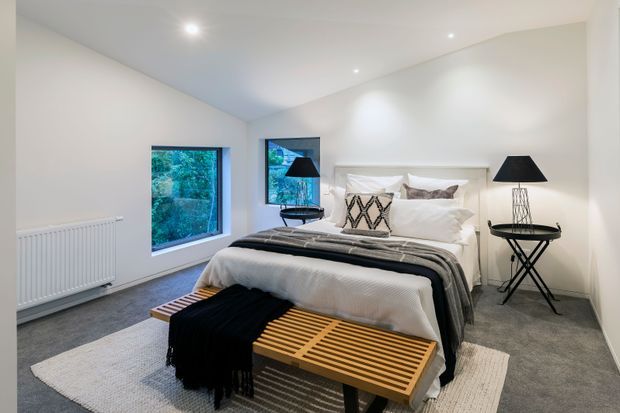 A bright, contemporary bedroom with a white upholstered bed, layered white and grey bedding, a patterned accent cushion, black bedside tables and lamps, and a slatted timber bench at the foot of the bed. The room has white walls and ceiling, grey carpet, recessed downlights, large windows overlooking greenery, and a light neutral rug.