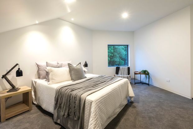 A bright, minimalist bedroom with a large upholstered bed dressed in layered white and grey bedding, a textured grey throw, and neutral cushions. The room has white walls and ceiling, recessed downlights, grey carpet, simple timber bedside furniture, black task lamps, a small black metal side table, and a window overlooking greenery.