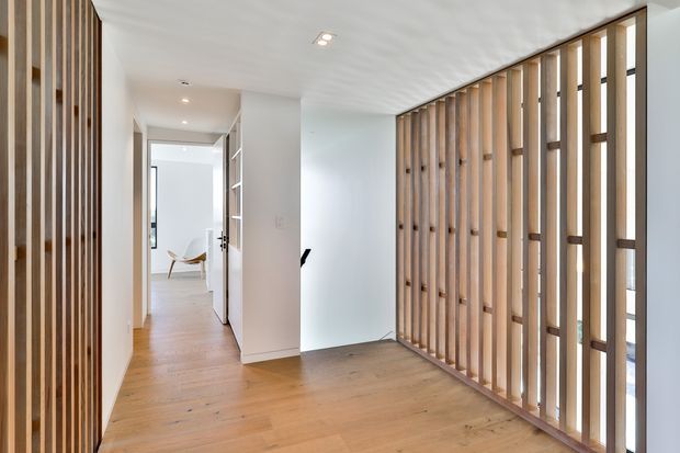 Bright contemporary interior hallway or entry space with warm timber flooring and prominent vertical timber slat screens along the right wall and left edge. White walls and ceiling, recessed downlights, and a glazed/open partition create an airy, minimalist transition space leading toward a furnished room.
