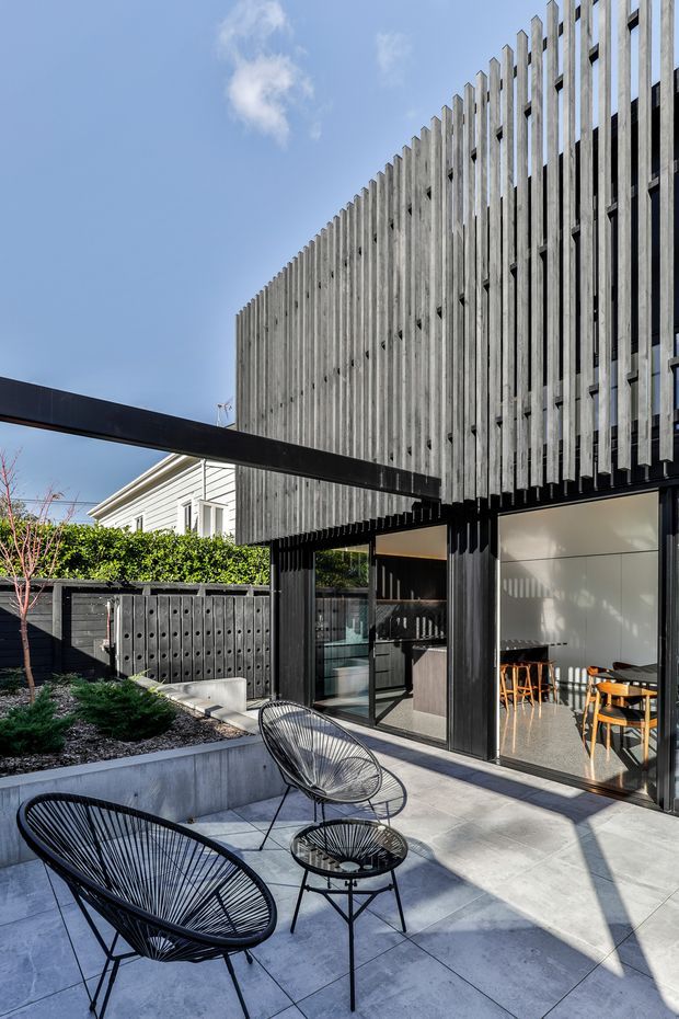 A contemporary courtyard patio beside a dark-framed glazed ground floor, with a striking upper facade of closely spaced vertical weathered timber fins. Pale concrete paving, black outdoor wire lounge chairs and a small table occupy the foreground. Large sliding glass doors reveal a bright dining area, while a neighbouring light weatherboard structure and landscaped planting are visible beyond.