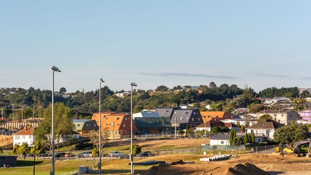 A wide exterior view across a suburban residential development, showing newly graded foreground land, construction fencing and materials, detached homes, low-rise buildings with pitched roofs, and a leafy hillside neighbourhood beyond. Tall site lighting poles and active earthworks indicate an ongoing construction or development site under clear daylight.