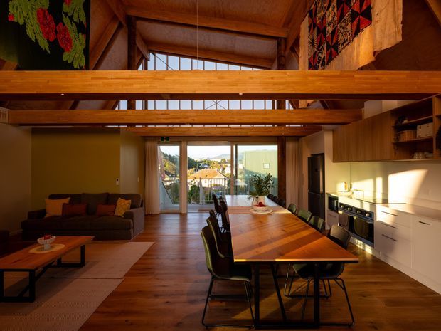 Open-plan timber-lined living, dining and kitchen interior with a dramatic double-height pitched roof and exposed timber beams. A long timber dining table occupies the centre, with a dark sectional sofa to the left and a compact white-and-timber kitchen with integrated appliances to the right. Full-height glazing opens toward an urban residential view, while colourful textile wall hangings add warmth and character.