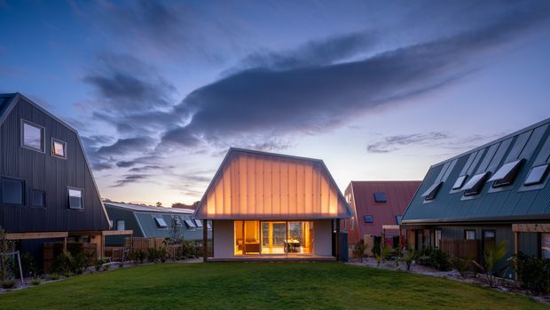 Twilight exterior view of a contemporary residential cluster arranged around a shared grassy courtyard. The homes feature steeply pitched gable roofs, dark vertical cladding, and prominent metal roof surfaces with skylights. A centrally positioned timber-clad dwelling glows warmly through its large ground-floor glazing, while similar houses extend to either side under a dramatic evening sky.