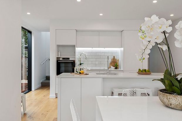 Bright, modern white kitchen and dining area with handleless cabinetry, a white subway-tile splashback, integrated oven, island or peninsula bench, and a white dining table. Light timber flooring and black-framed glazing add contrast, while large white orchid arrangements provide decorative accents. A hallway and stair are visible to the left.