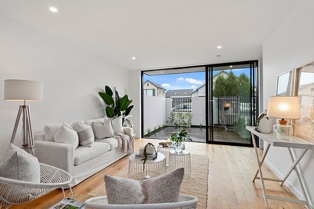 Bright contemporary living room with white walls, pale timber flooring, a light upholstered sofa and lounge chairs, woven rug and coffee tables. Full-height black-framed sliding glass doors open to a small balcony or terrace, with greenery and neighbouring homes visible beyond. A slim console, lamps and indoor plants complete the airy neutral interior.