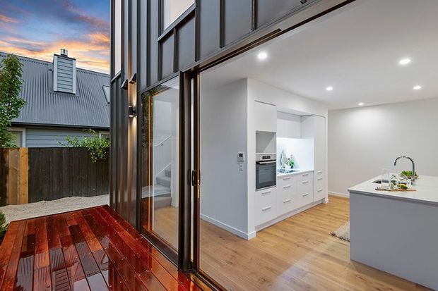 Contemporary open-plan kitchen viewed from a wet timber deck through large glazed doors. The bright white kitchen features integrated cabinetry, a central island with a sink, recessed ceiling lighting, and light timber flooring. Dark metal-framed glazing and vertical exterior cladding contrast with the warm deck and neighbouring pitched roof visible outside.