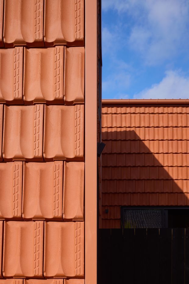 Close-up architectural detail of a terracotta tiled roof and facade, showing repeating profiled roof tiles, a vertical terracotta-clad edge, and a darker tiled building section against a partly cloudy blue sky. Strong sunlight creates deep shadows across the roof surface.