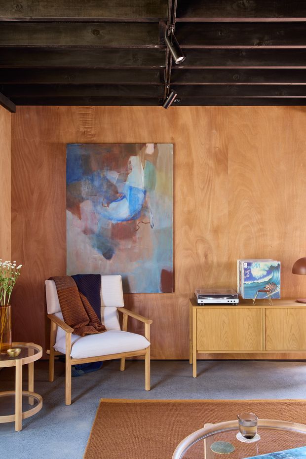 Warm, richly layered living room vignette featuring full-height timber wall panelling, exposed dark timber ceiling beams with black track lighting, a mid-century style upholstered armchair, a timber media sideboard, turntable, artwork, and a round glass coffee table. The room has a refined vintage-modern character with earthy textiles, blue-grey carpet, and warm wood tones.