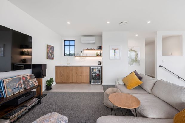 Bright contemporary open-plan living room with a compact built-in bar or kitchenette at the rear. The space features white walls and ceiling, recessed lighting, timber cabinetry, a dark integrated media wall, grey carpet, a large light-grey sectional sofa, and a round timber coffee table. Artwork, shelving, and a yellow accent cushion add warmth and colour. A stair or hallway opening is visible to the right.