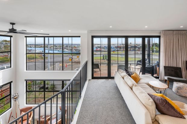 Bright contemporary open-plan living area on an upper landing, featuring expansive black-framed glazing, a carpeted floor, a large light sectional sofa, and a black metal balustrade overlooking a lower level. The windows and glazed doors provide views toward a marina, sports fields, and surrounding urban development. A ceiling fan, recessed downlights, neutral curtains, and warm accent cushions complete the space.
