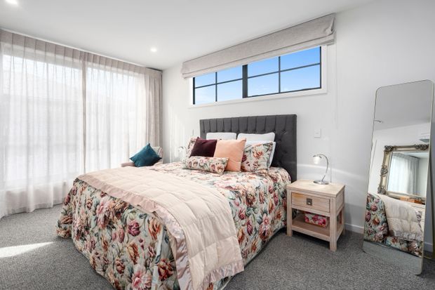 Bright, well-appointed bedroom with a tufted dark upholstered bedhead, floral patterned bedding, pastel cushions, bedside table and lamp, full-length mirror, sheer curtains, and a high horizontal multi-pane window. The room has light painted walls, recessed ceiling lights, and grey carpet.