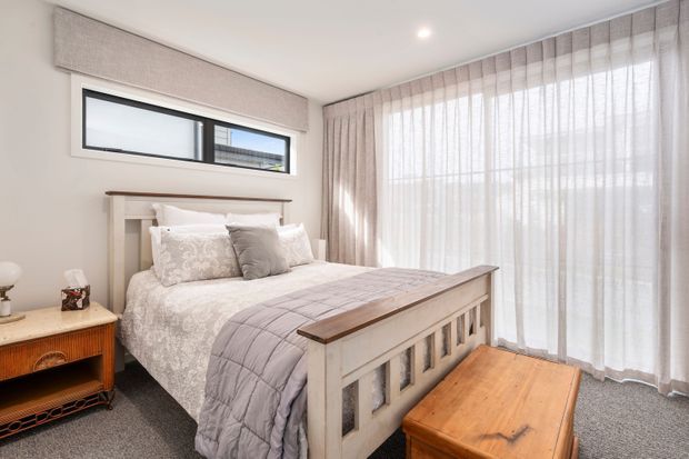 Bright, softly styled bedroom with a white-painted timber bed featuring a paneled footboard and a simple matching headboard. The bed is dressed in white and pale grey bedding with patterned pillows and a quilt. Full-height sheer curtains cover large glazing on the right, while a narrow horizontal window sits above the headboard. A warm timber bedside table and timber trunk add contrast against light walls and grey carpet.
