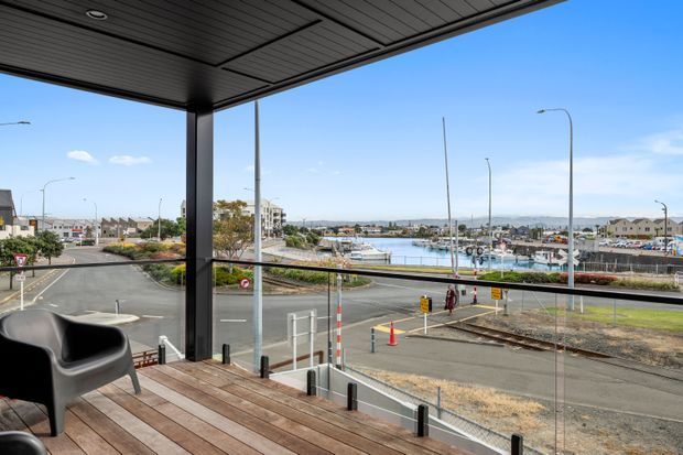 A covered balcony or terrace overlooking a marina and surrounding urban streets. The space has a dark slatted ceiling, slender dark structural posts, full-height clear glazing or balustrade panels, and a timber deck with a black lounge chair. Bright daylight and blue sky create an open waterfront outlook.