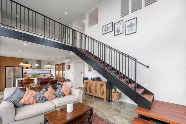 Bright contemporary open-plan living area with a large sectional sofa, patterned cushions, timber coffee table, and a visible kitchen and dining area beyond. A striking black metal staircase with timber treads rises to a mezzanine level, complemented by timber flooring and cabinetry, white walls, framed botanical artwork, and indoor plants.