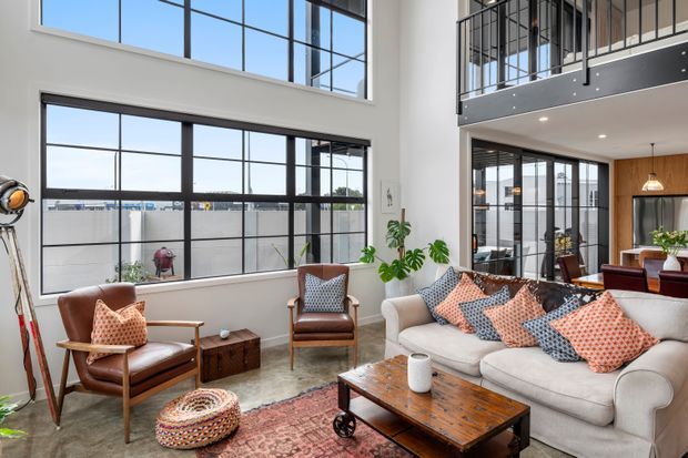 Bright, spacious contemporary living room with a large cream sectional sofa, leather armchairs, patterned cushions, a substantial timber coffee table, and a vintage-style rug. Floor-to-ceiling black-framed windows and a double-height void with an upper-level balcony create strong industrial loft character, while pendant lighting, indoor plants, and eclectic furnishings add warmth.