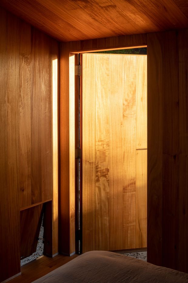 A warm, intimate interior view from a bedroom toward a partially open timber door. The walls, ceiling, door, and surrounding built-in surfaces are extensively clad in rich, vertically grained dark wood, with strong sunlight entering through the doorway and illuminating the lighter timber door panel. The lower foreground shows the edge of a bed, confirming the bedroom setting. A narrow view of gravel and greenery is visible outside.