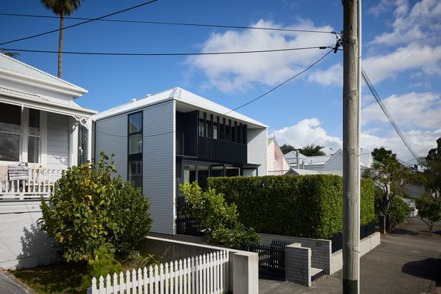 Street-facing view of a compact contemporary two-storey house in a suburban residential setting. The home has light horizontal cladding, a low-pitched hipped roof, dark vertical screening and window frames, a hedge-lined frontage, and a small paved entry area. A traditional weatherboard house with ornate trim and a white picket fence sits adjacent.