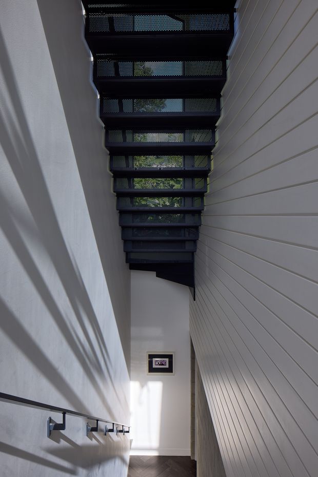 A narrow, tall interior passage or stairway-like circulation space with white horizontal wall panelling on the right, a smooth white wall on the left, dark timber flooring, and a series of black perforated overhead screens or stair treads creating strong linear shadows. Minimal wall-mounted hooks or brackets run along the lower left wall, with a small framed artwork at the far end. Natural light filters through the overhead structure and greenery is visible beyond.
