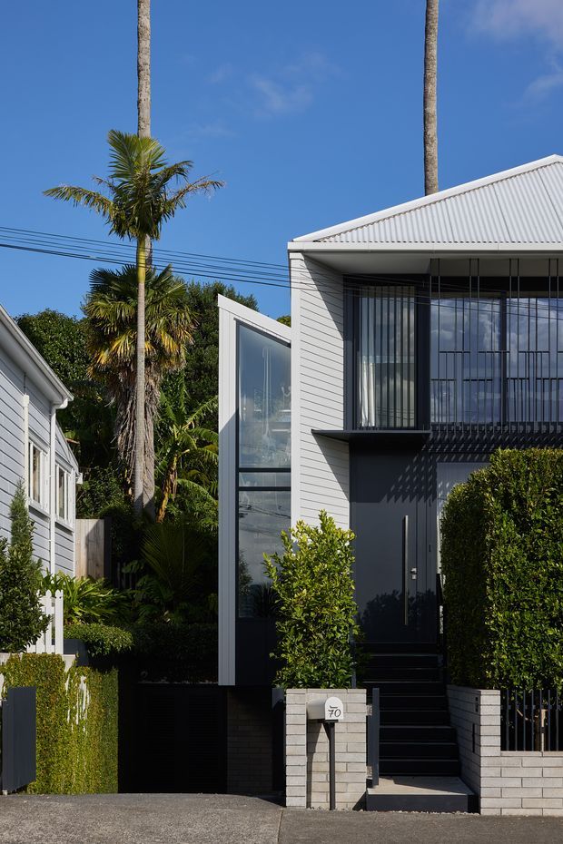Street-facing view of a narrow contemporary multi-storey residence with a white horizontal-clad facade, steep corrugated metal gable roof, tall glazed entry volume, dark recessed front door, upper-level balcony or screened glazing, and pale masonry boundary walls. Dense subtropical planting and neighbouring homes frame the property.