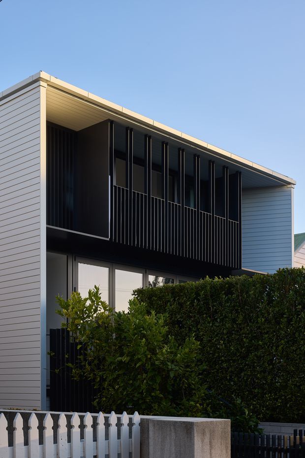 Exterior view of a contemporary two-storey house with pale horizontal cladding, a broad low-pitched roof overhang, dark recessed upper-level balcony or screening, and tall glazed openings below. Dense hedging and a front fence partially obscure the lower facade.