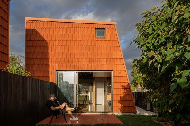 Exterior view of a compact contemporary dwelling with a steeply sloped, terracotta-coloured tiled facade and roof-like wall form. Large glazed sliding doors open onto a small paved patio, with a glimpse of the interior, while dark fencing and mature planting frame the scene under dramatic cloudy skies.